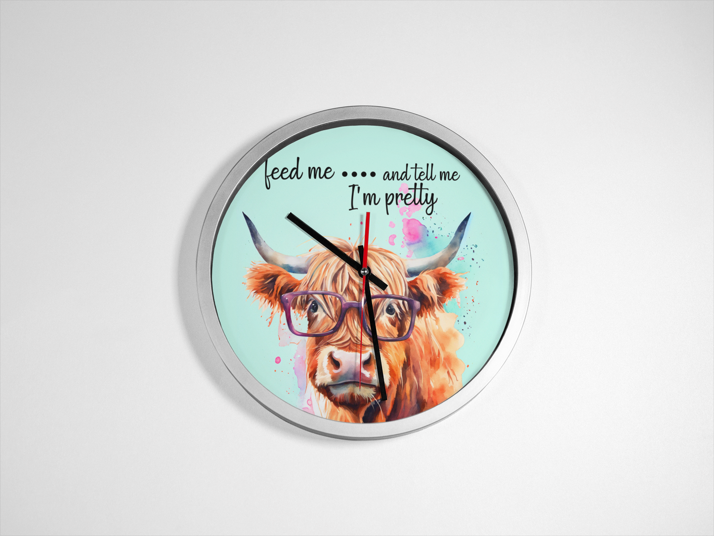 A wall clock featuring an illustration of a Highland cow with glasses, with a caption 'feed me... and tell me I'm pretty'.